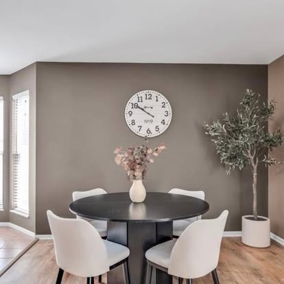Staged dining area featuring taupe accent wall, round dark espresso pedestal table, off-white upholstered bucket chairs, and potted olive tree.